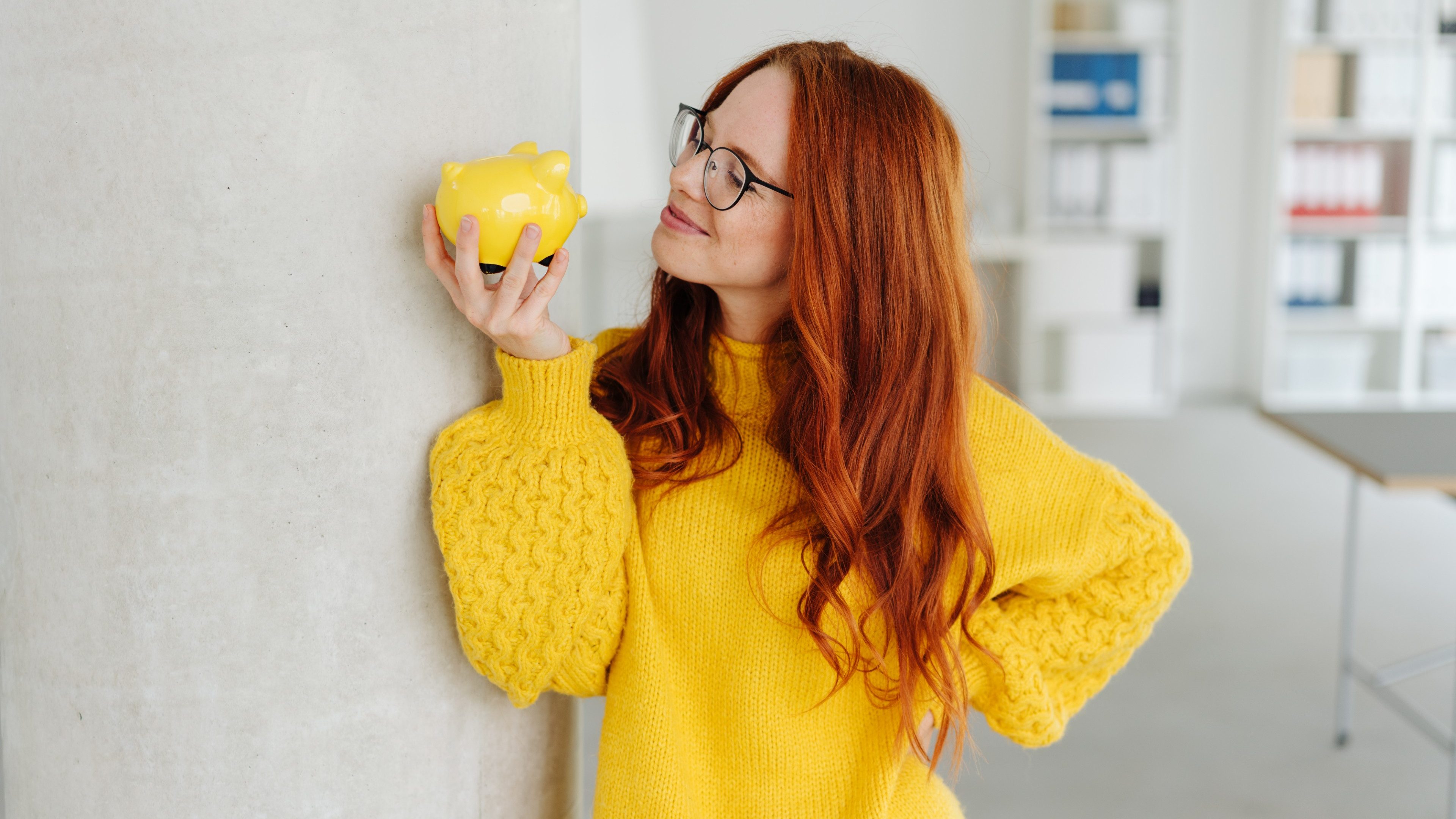 Smiling young businesswoman contemplating a piggy bank as she plans what to do with her savings in a bright high key office