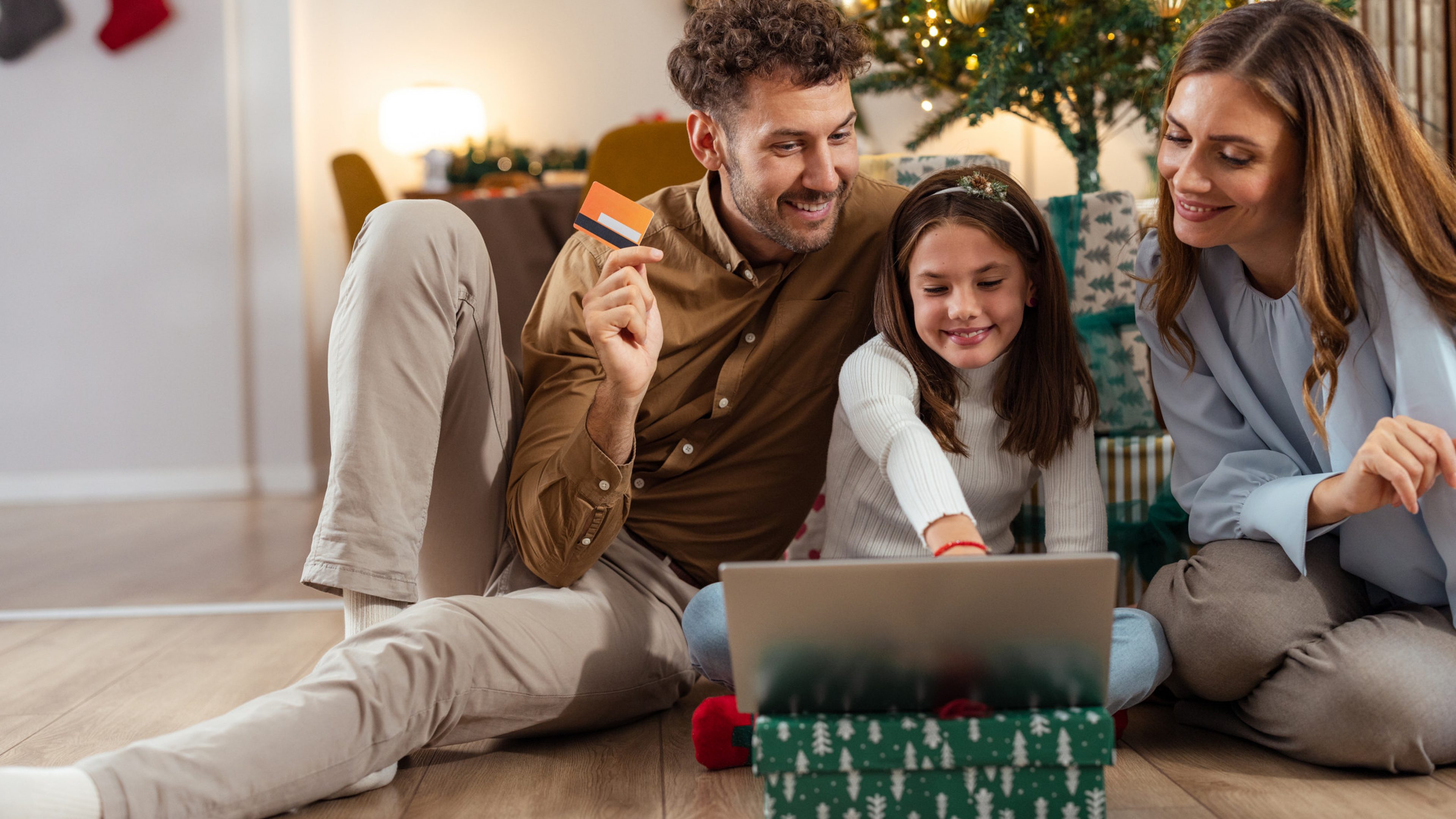 A joyful family of three sits on the floor during Christmas, with the father holding a credit card and looking at the laptop.  A daughter happily sits between parents, enjoying the cozy, festive atmosphere.