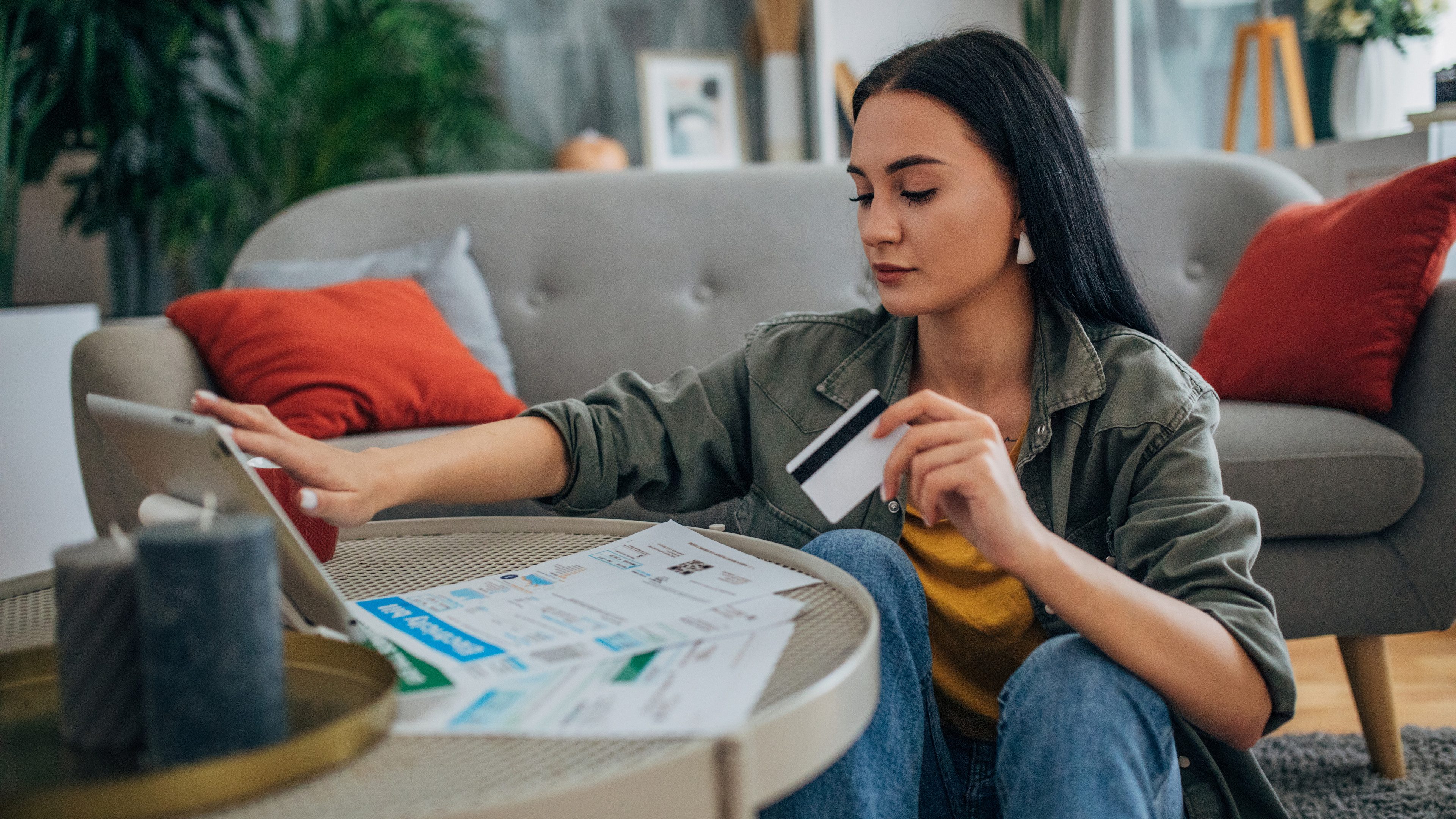 A woman shops online using her tablet while holding a credit card. She is seated in a chic living room, featuring a gray sofa, red cushions, and modern decor, creating an elegant atmosphere.