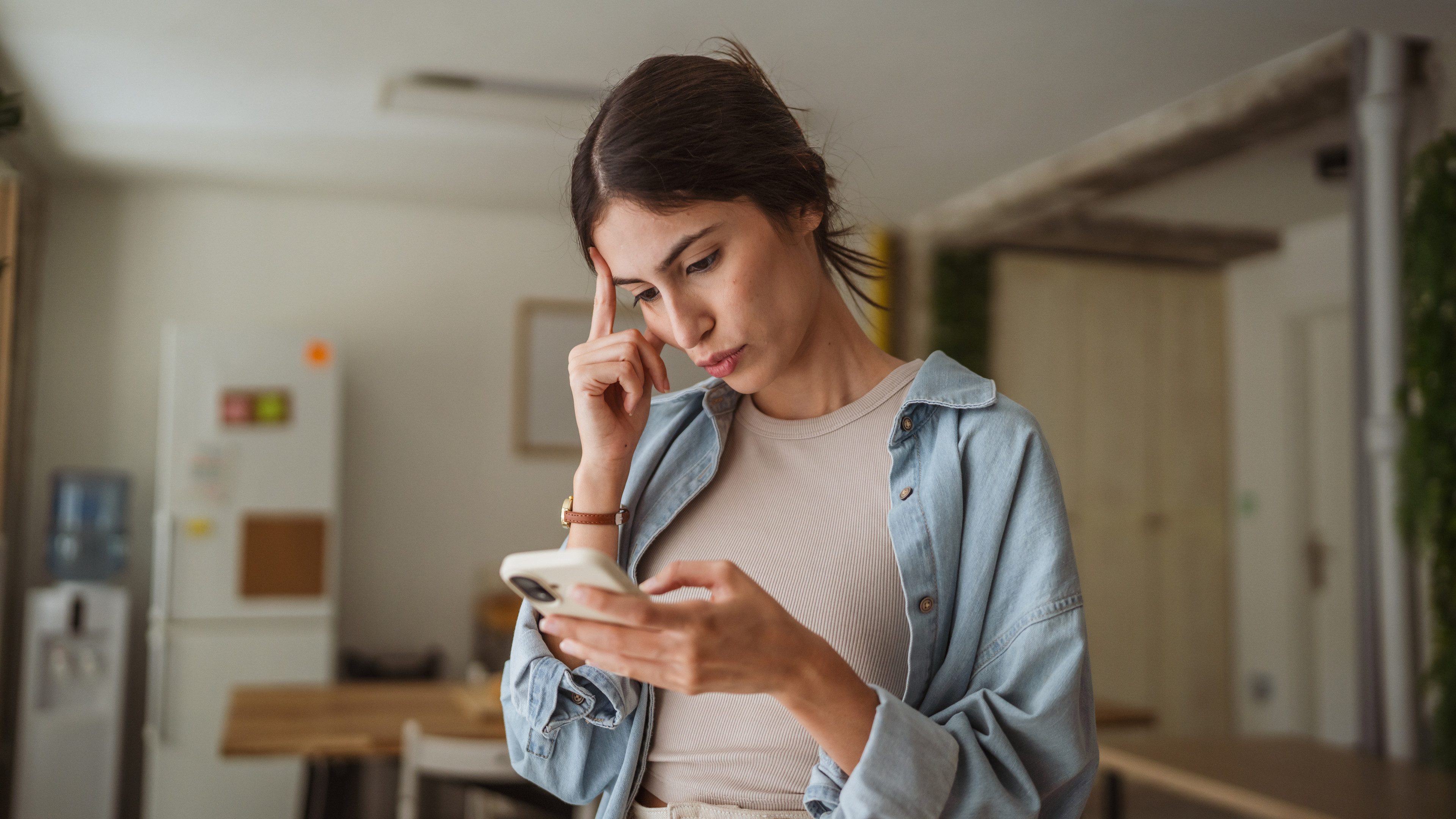 Young businesswoman is having problems while working from home and reading a message on her smart phone, touching her forehead with a thoughtful expression