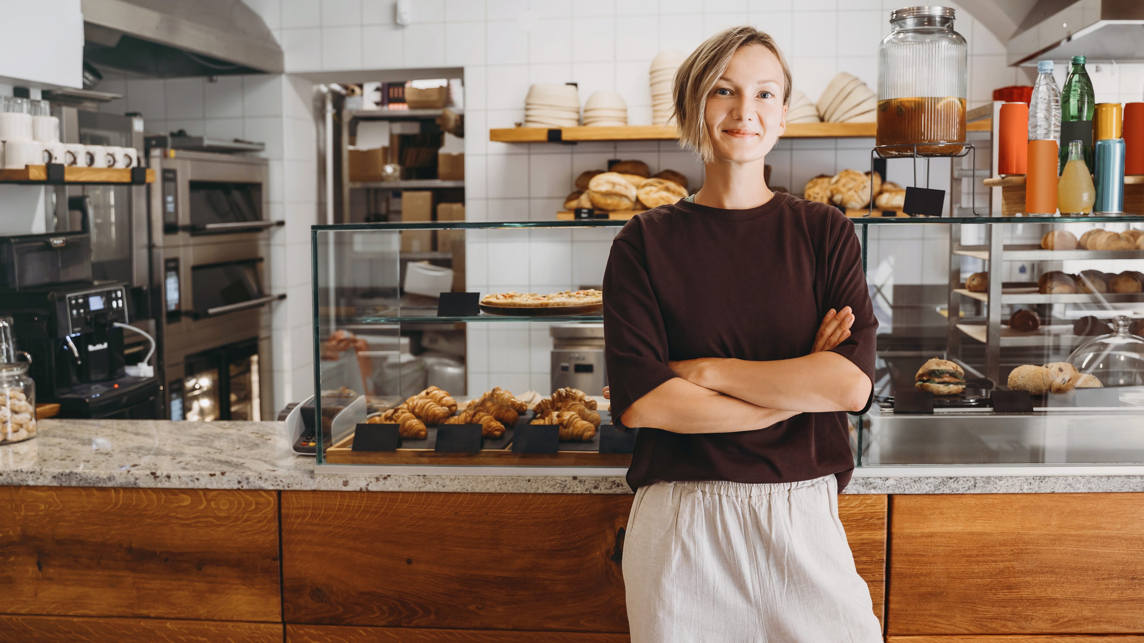 Portrait of smiling young woman entrepreneur standing at the counter of her bakery and coffee shop. Local small business owner indoors. Female in cafe near showcase with fresh croissants and bread.