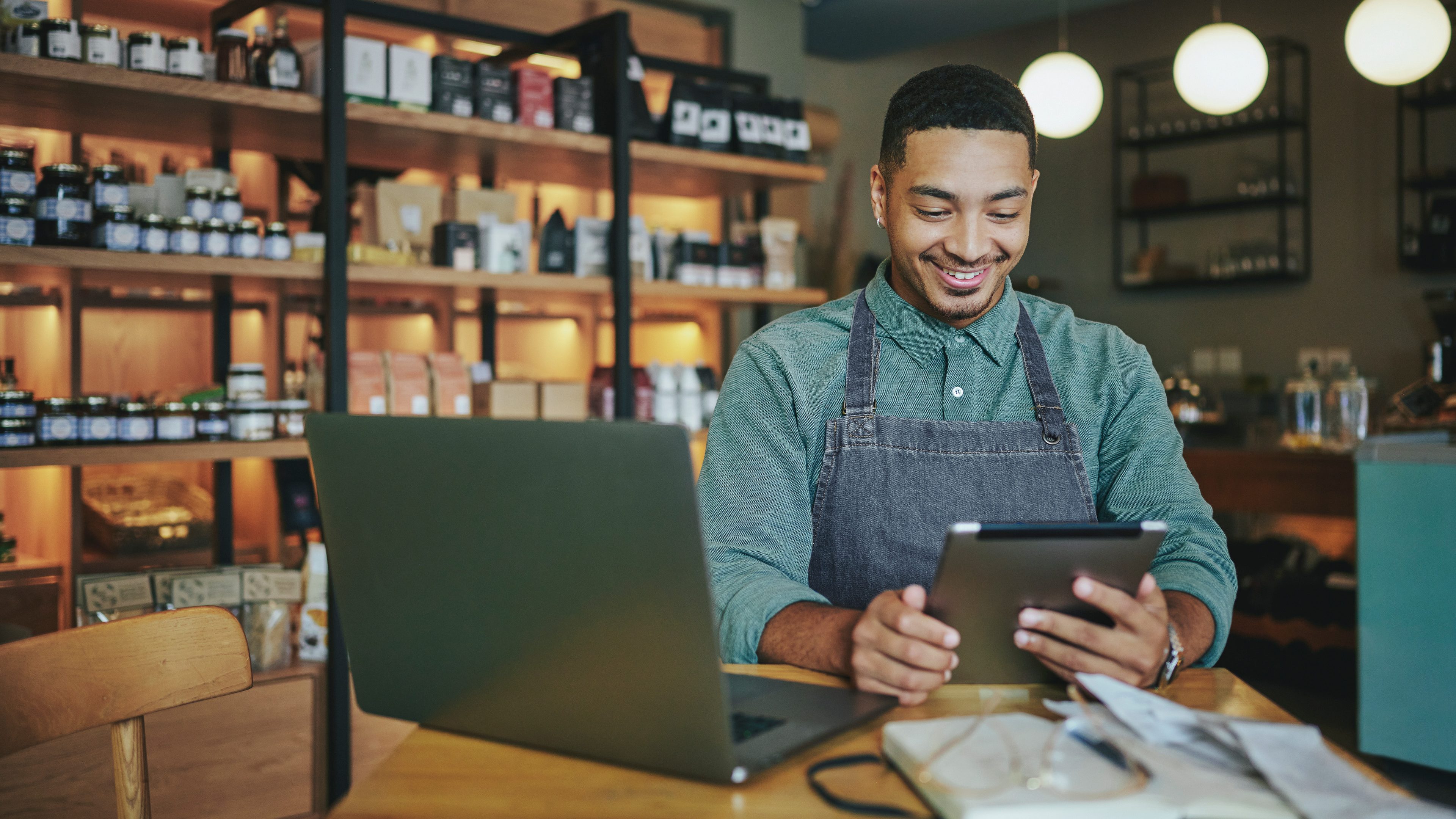Smiling young male deli owner working a tablet and a laptop while sitting at a table in his artisanal shop