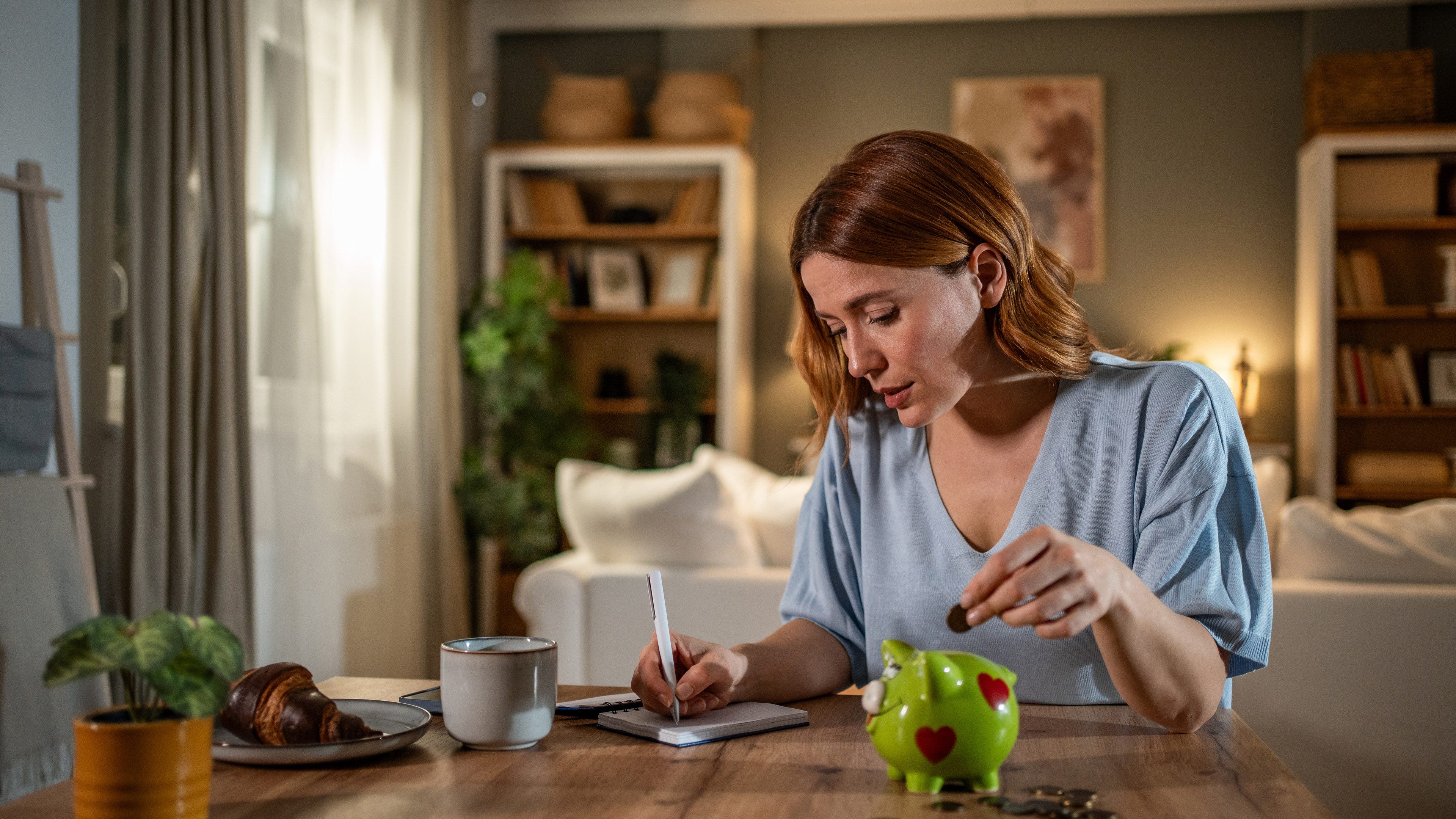Young woman carefully managing her home finances, putting coins in a piggy bank and taking notes on expenses and savings in her living room