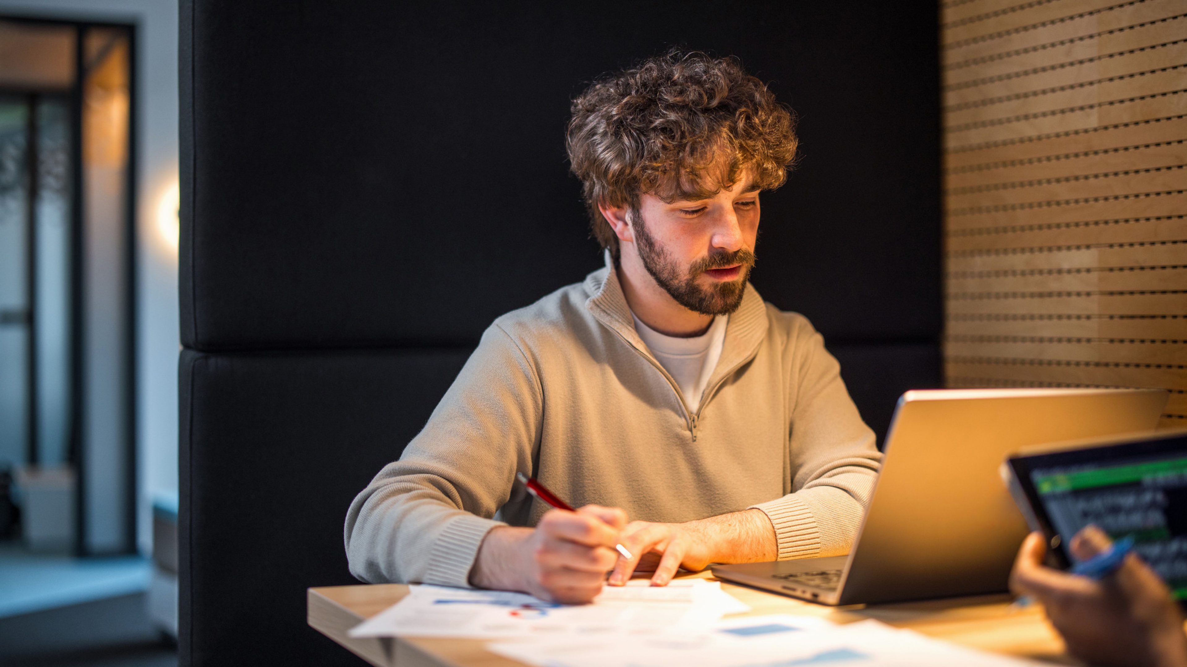 A professional Hispanic man engaged in examining financial charts on paper while using a laptop in a modern office setting.