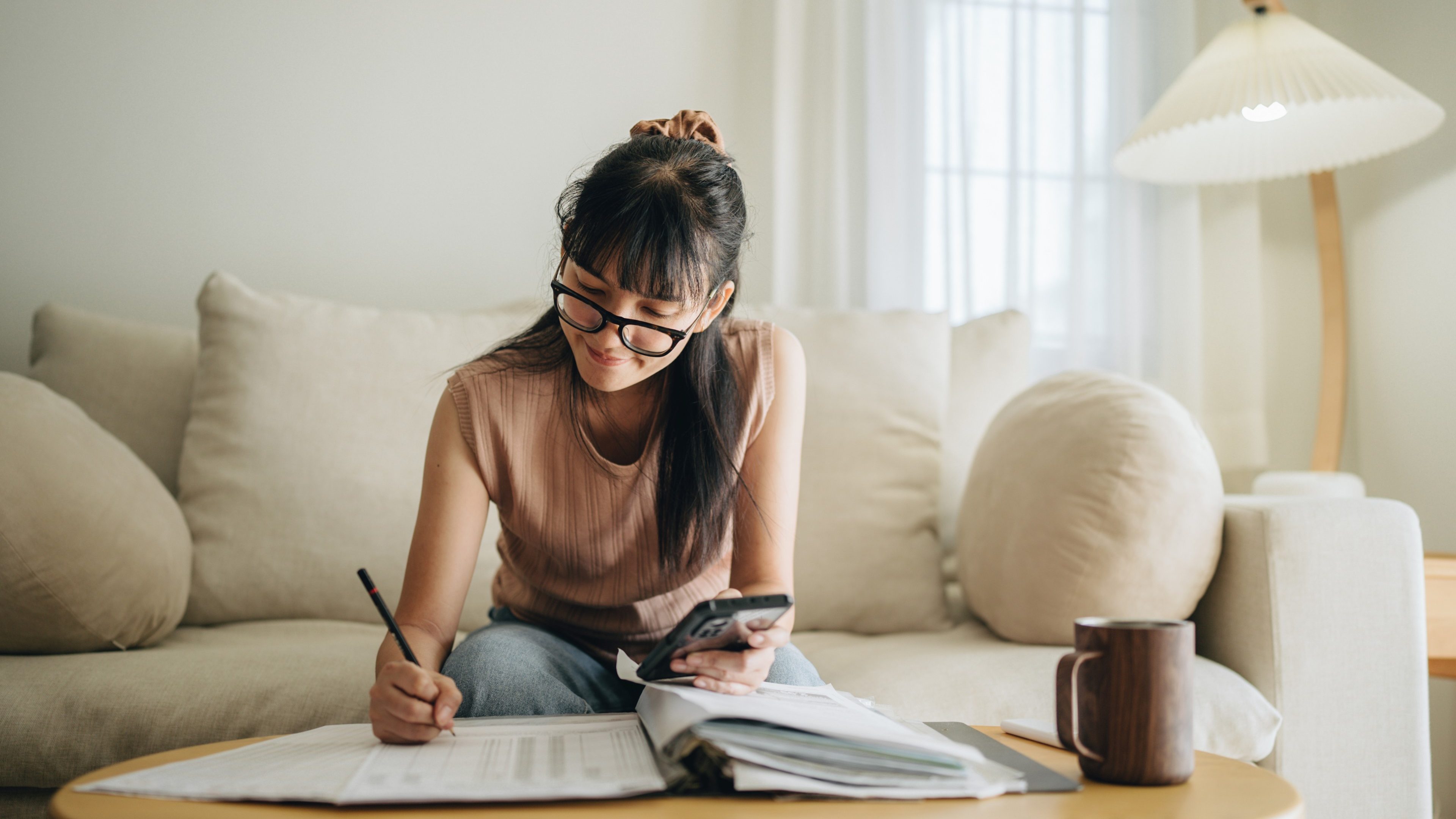 A woman sits at her living room with smartphone and financial reports doing her monthly budget.