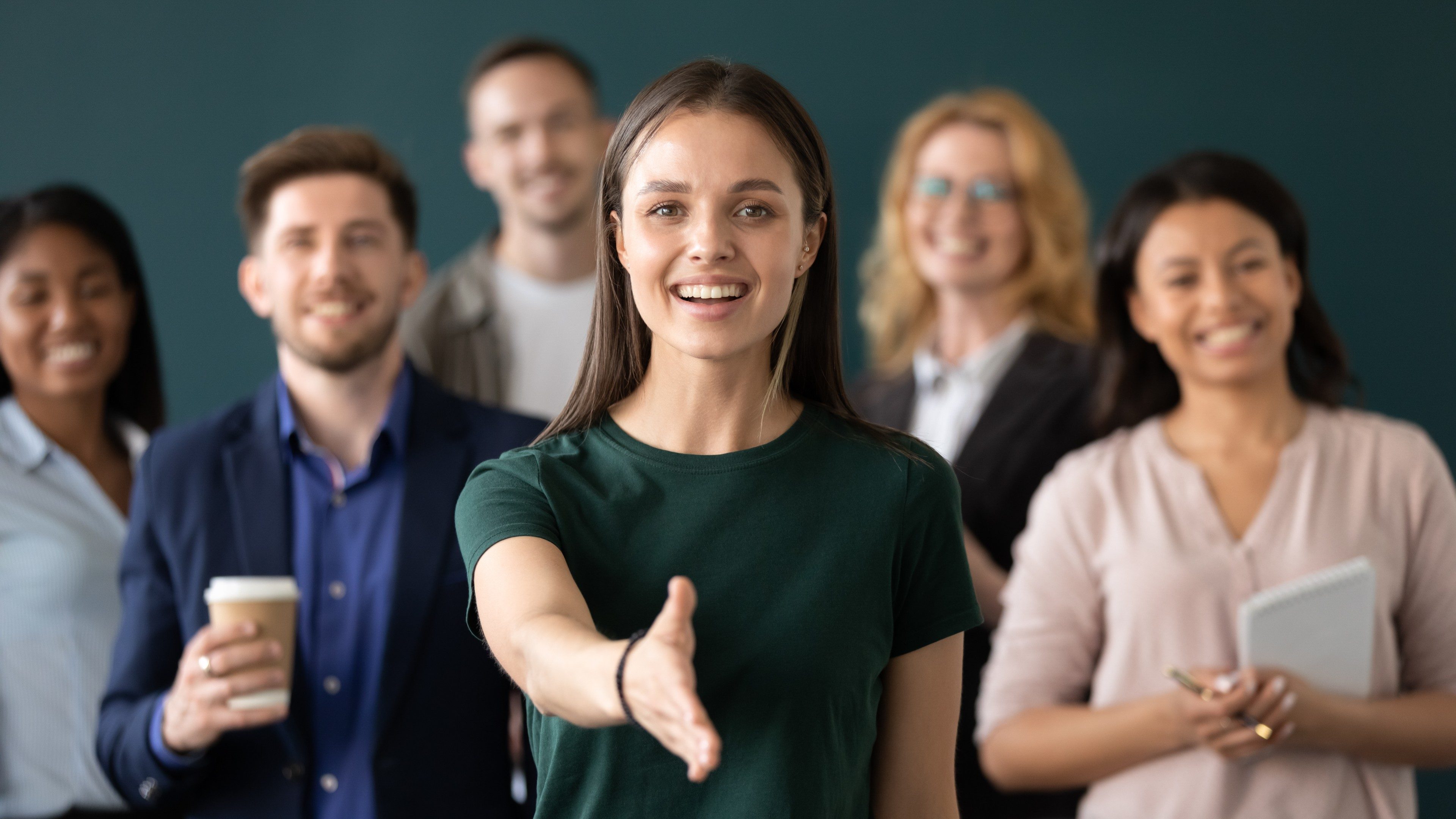 Friendly young company woman representative holds out her hand for handshake welcoming customer smiling looking at camera posing together with diverse colleagues, sales manager greeting client concept