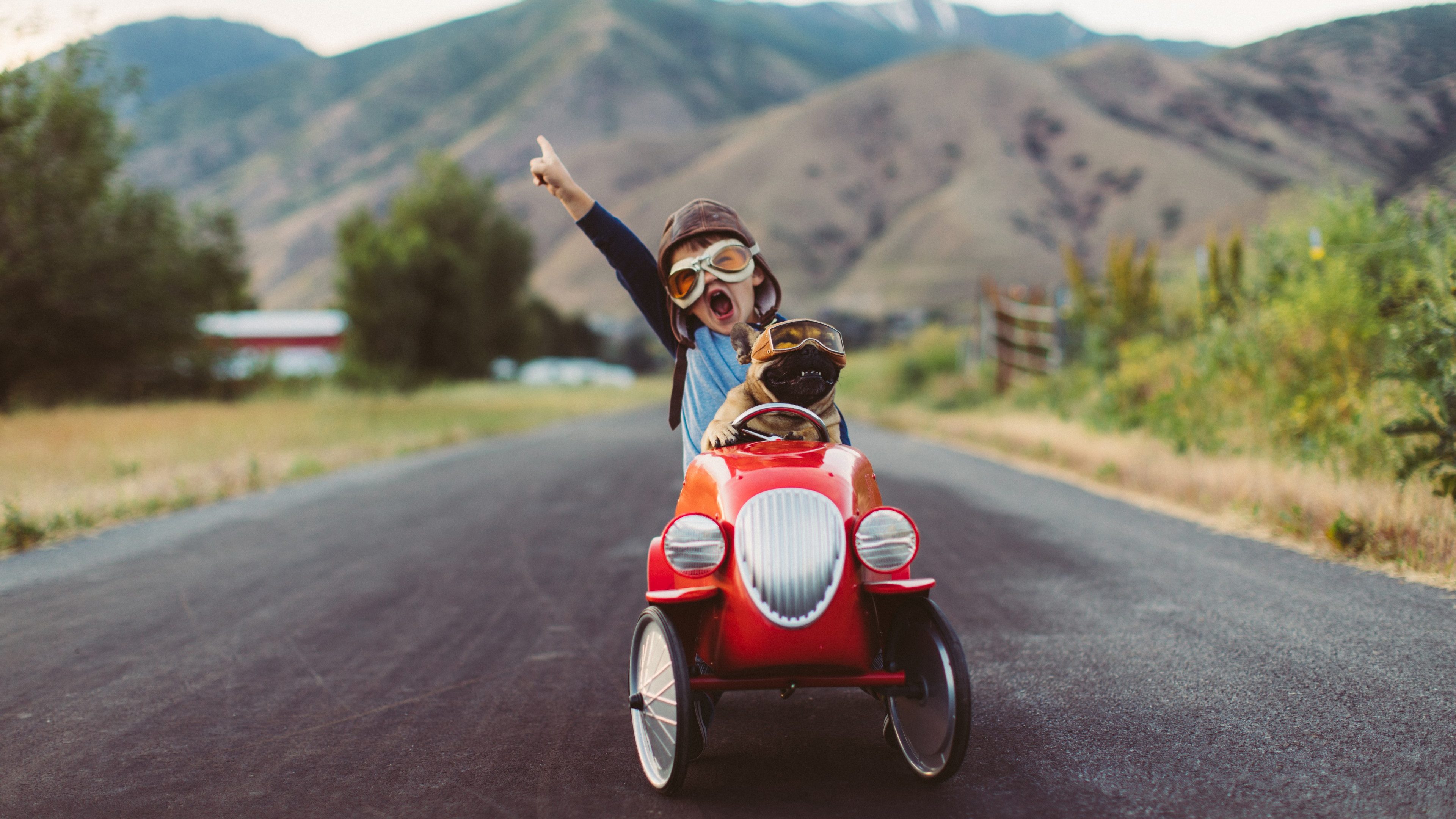 A young boy with flying goggles and flight cap races a red toy car with his pet and best friend French Bulldog along a small road in Utah, USA. Sometimes a road trip journey with your best friend and some fresh air in your face is the best medicine for the soul.