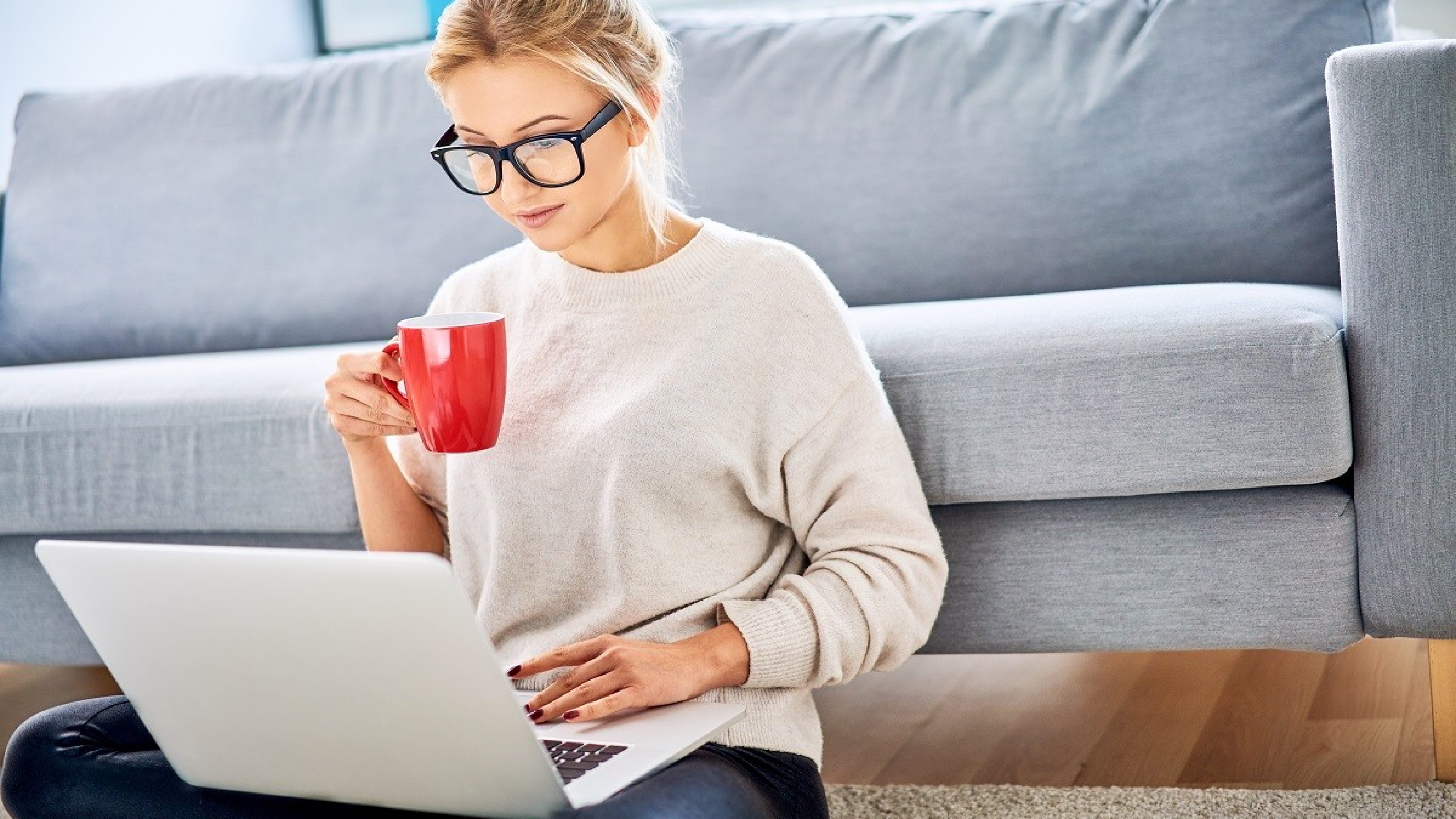 Young woman having coffee while using laptop and sitting on floor at home