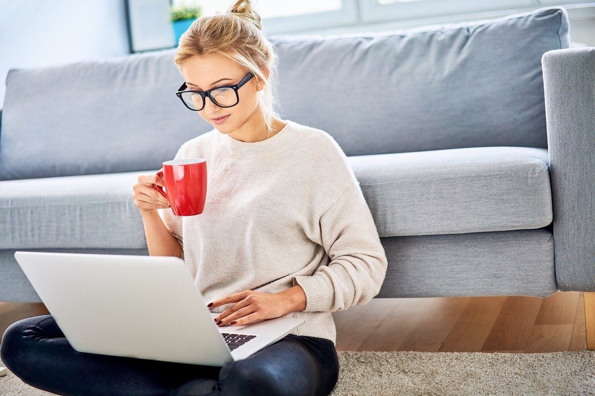 Young woman having coffee while using laptop and sitting on floor at home